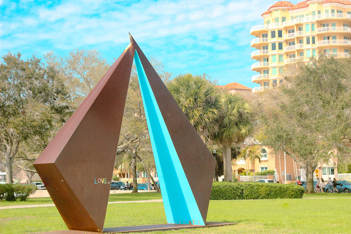 A park, featuring a large brown and blue metal sculpture with LOVE written on it in chalk. Behind it, a stucco apartment building, palm trees, and a lightly cloudy sky.