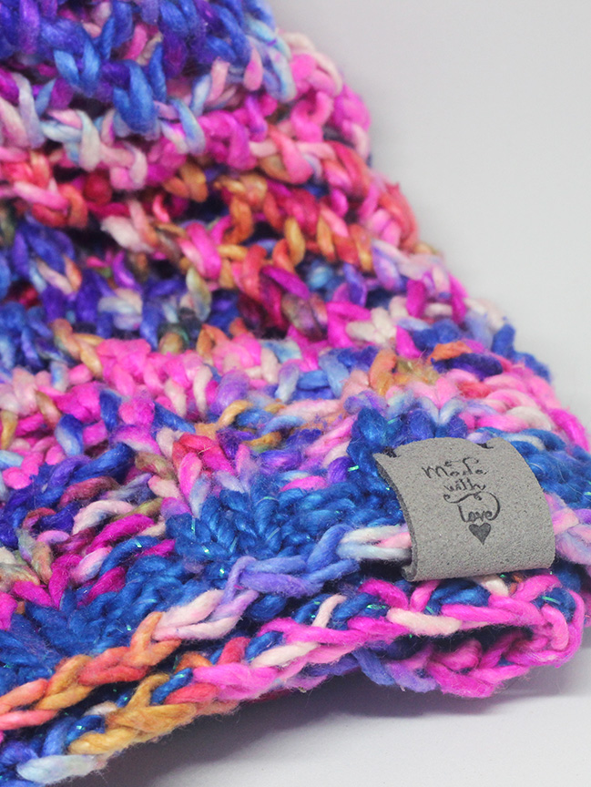 a close up photo of a pink, purple, blue, and orange hand-knit hat. A grey faux-suede tag on the brim says made with love.